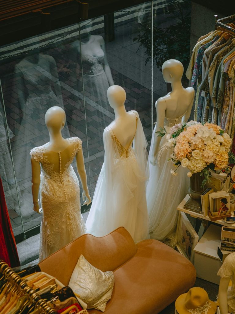 From a high-angle view, three mannequins showcase elegant wedding gowns in a charming bridal boutique within Hong Kongs historic Western Market. The dresses feature intricate lace and delicate fabrics, their forms reflected in a large window that adds a dreamy quality to the scene. A plush, dusty-rose sofa and racks of vintage-style garments create a cozy, curated atmosphere.